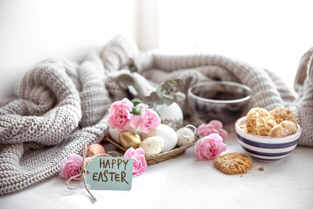 Натюрморт с чаем, печеньем, яйцами, цветами и надписью Happy Easter на открытке.
