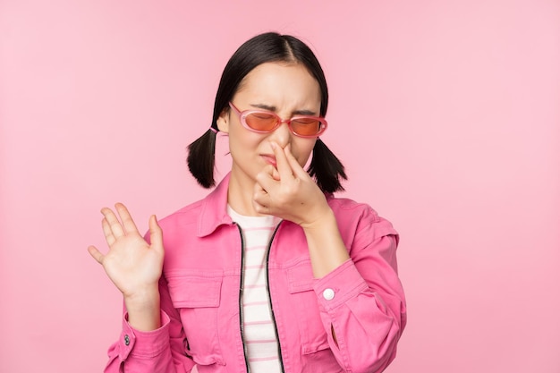 무료 사진 선글라스와 세련된 옷을 입은 냄새 나는 아시아 소녀는 분홍색 배경 위에 서있는 혐오감 싫어서 코를 닫습니다