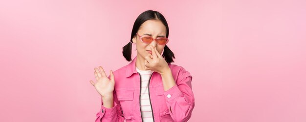 Stinky Asian girl in sunglasses and stylish outfit shuts her nose from disgust dislike bad smell standing over pink background
