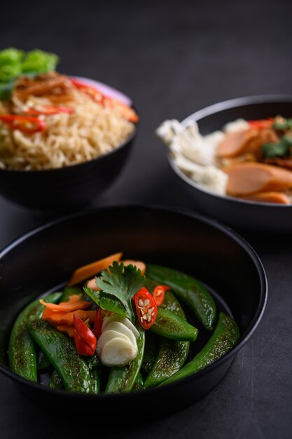 Stir-fried peas in a frying pan and noodles spicy in the bowl with ingredients on black cement surface.