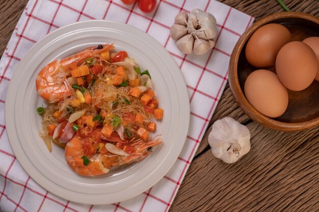 stir-fried prawns with glass noodles in a white plate placed on a cloth with eggs and garlic.