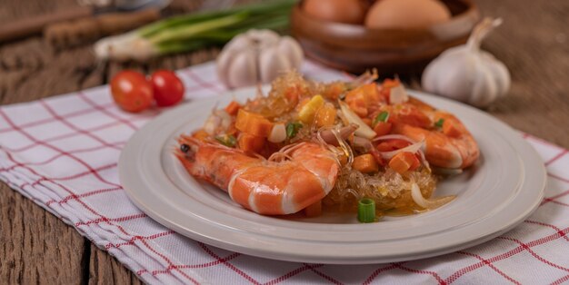 Stir-fried prawns with glass noodles in a white plate placed on a cloth with eggs and garlic.