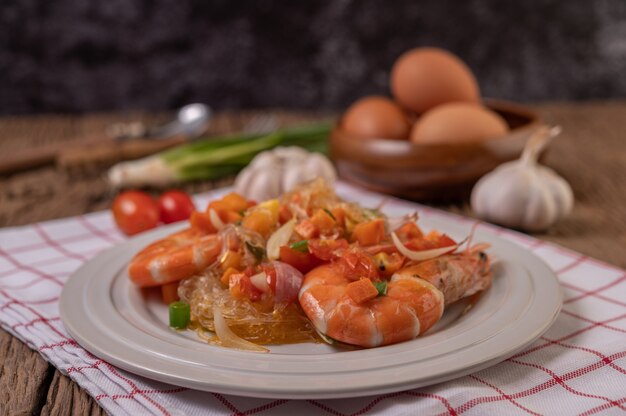 stir-fried prawns with glass noodles in a white plate placed on a cloth with eggs and garlic.