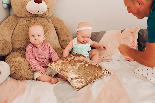 Stock photo of adorable kids - sister and brother - sitting on bed with big teddy bear. Dad playing with two cute babies on the bed.