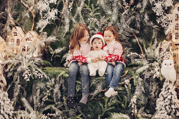 Stock photo of adorable little baby boy in Santa hat embraced by his elder sisters in red and white sweaters with winter pattern. Children with baby sitting in Christmas interior.