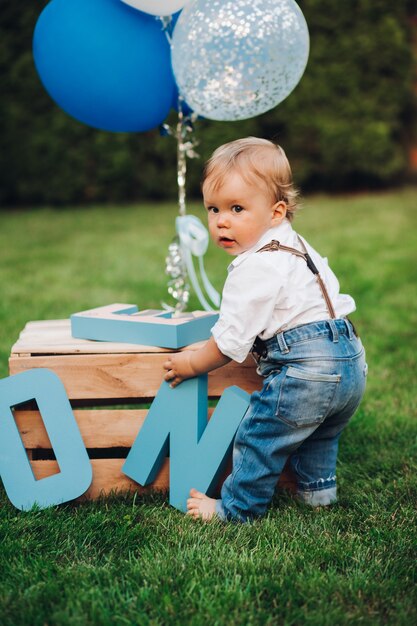 Stock photo of an adorable little boy in jeans, shirt and suspenders playing with birthday decorations on the lawn in the backyard. Summer day out. Birthday concept. Air balloons and wooden letters.