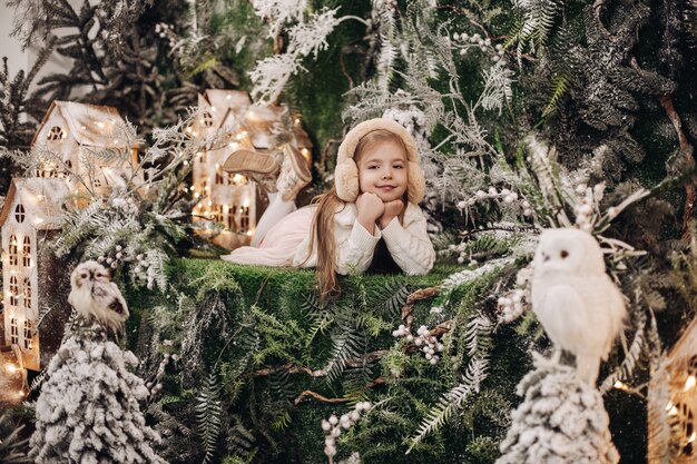 Stock photo of adorable little Caucasian child in beige earmuffs laying with chin on hands surrounded by Christmas decorations and smiling at camera. Winter wonderland concept.