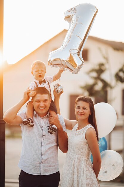 Stock photo of a beautiful Caucasian family with baby son holding inflatable number one. Outdoors. Fair weather and sunlight.