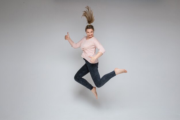 Stock photo of cheerful carefree blonde woman jumping in mid-air with thumbs up on white background. Jumping woman smiling at camera holding thumbs up.