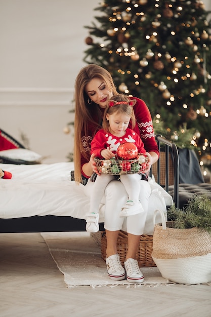 Stock photo of loving mother in green dress giving her little daughter in pyjama dress a Christmas present. They are next to beautifully decorated Christmas tree under snowfall.