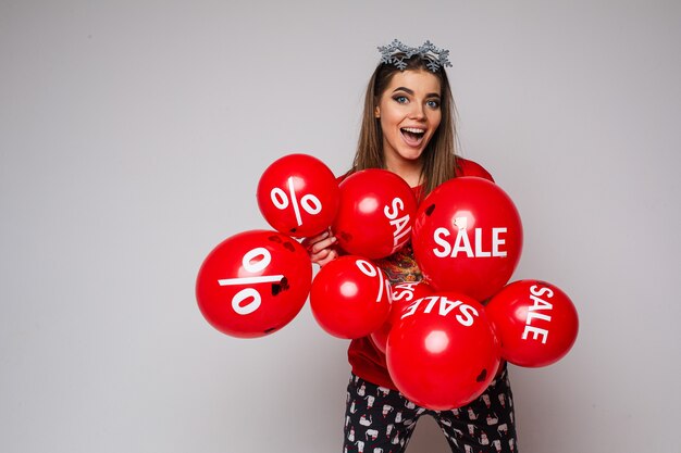 無料写真 頭にスノーフレークの派手な眼鏡をかけ、販売と割引のステッカーと赤い気球の束を保持しているパジャマで興奮したブルネットの女性のストックフォト。