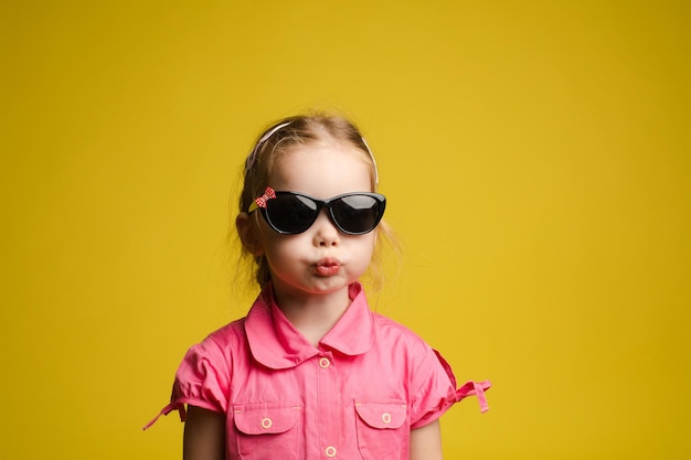 Free photo stock photo portrait of adorable little kid in fashionable black sunglasses pouting her lips funnily on yellow background isolate