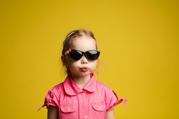 Фотопортрет очаровательного маленького ребенка в модных черных солнцезащитных очках, забавно надувающих губы на желтом фоне Изолировать