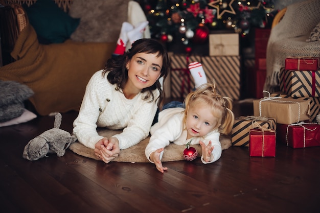 Stock photo portrait of attractive young adult mother with little blonde daughter laying on the wooden floor over pillow with wrapped Christmas presents. Girl is playing with red Christmas ball.