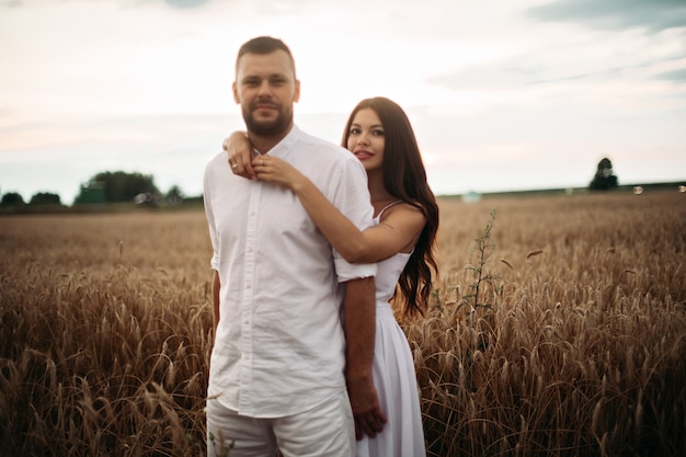 Фото портрет бородатого парня, обнимающего свою великолепную подругу в белой одежде, обнимая в пшеничном поле. Красивое пшеничное поле на заднем плане.