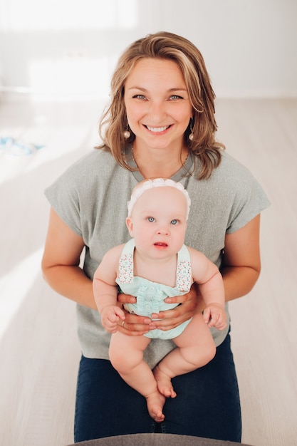 Stock photo portrait of a beautiful smiling mother in grey t-shirt and jeans holding her lovely little baby daughter in her hands and looking at camera happily...