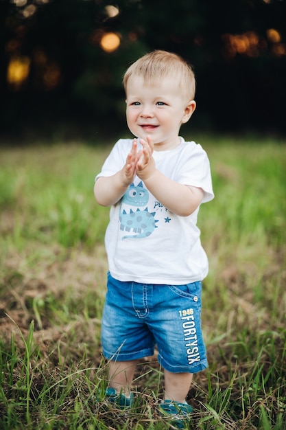Stock photo portrait in t-shirt and denim shorts clapping hands and smiling standing on green lawn in park. Bokeh background. Cheerful baby boy standing on grass with hands clapping.