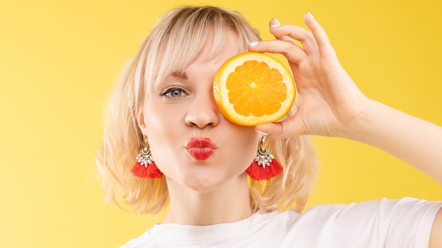 Stock photo of positive blonde young woman in white t-shirt with halved orange holding it in front of her eye and pouting lips at camera. Isolate on yellow background. Summer concept.