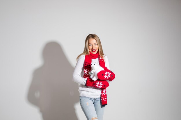 Stock photo of pretty blonde Caucasian woman in mittens and scarf and white sweater holding cute white toy bear and smiling.