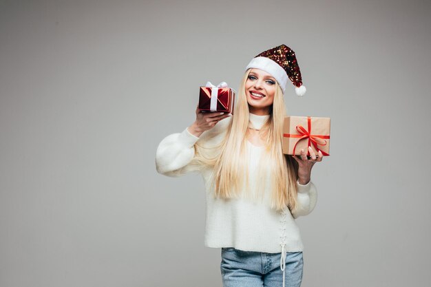 Stock photo of pretty blonde girl in sparkling Santa hat and sweater holding two wrapped christmas gifts in both hands. Choice concept.