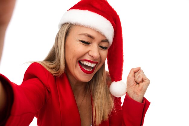 Stock photo of pretty jovial blonde Caucasian woman in red Santa hat and jacket squinting her eye with open mouth at camera making selfie. Studio shot isolate on white.