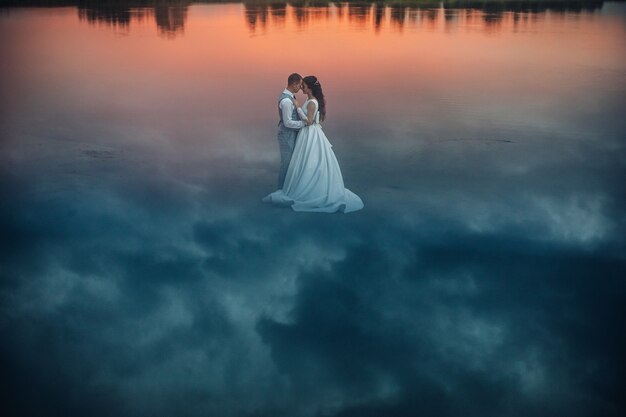 Фото запаса романтической невесты в свадебном платье и жениха в костюме, обнимающего лицом к лицу, стоя на мокром песке с отражением неба на нем. Облака отражаются от земли, создавая фантастический вид.
