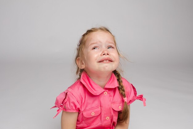 Stock photo of sweet little girl with braids in pink dress crying while sitting on the floor with bare feet She is looking at the camera while sobbing