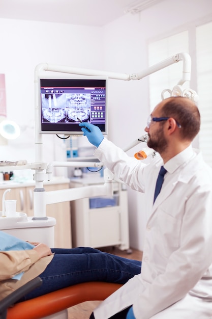 Stomatolog explaining dental treatment to senior woman during examination looking at x-ray. medical teeth care taker pointing at patient radiography on screen sitting on chair.