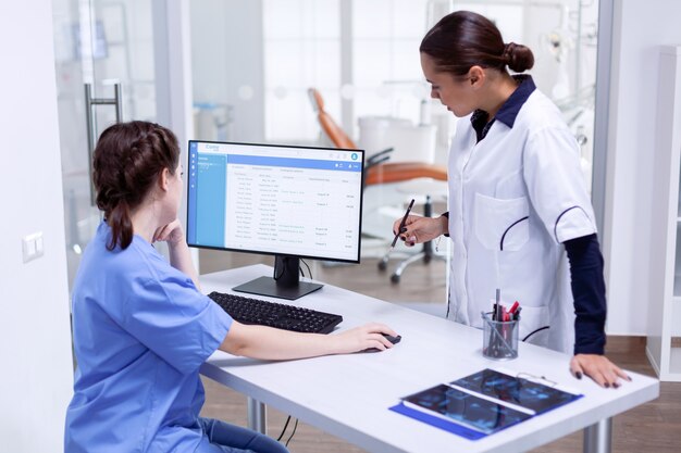 Stomatolog and nurse in tooth clinic checking patient appointment looking at computer monitor. Stomatology assistant and teeth doctor discussing in reception of dental office