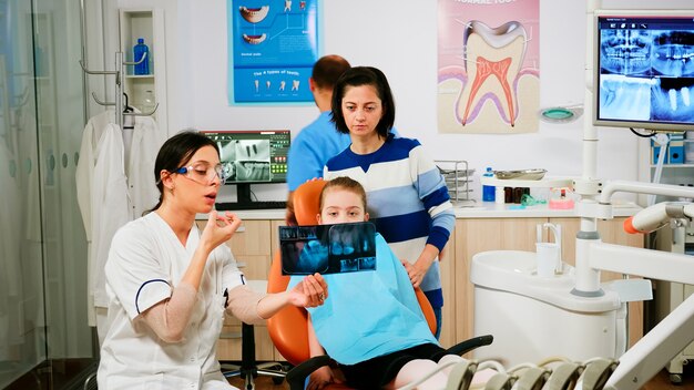 Stomatologist explaining dental treatment holding radiography pointing on affected teeth while man assistant preparing sterilized tools for surgery. Doctor and nurse working in stomatological unit