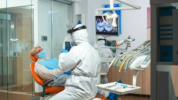 現代​の​クリニック​で​の​コロナウイルスパンデミック中​の​歯​の​問題​を​調べる​患者​の​歯科用​x線​写真​を​求める​防護服​を​着た​口腔病​専門医​。​つなぎ服​、​フェイス​シールド​、​マスク​、​手袋​を​着用した​医療​チーム​。