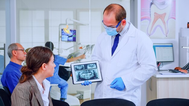 Stomatologist pointing on digital screen explaining x-ray to woman sitting on chair in waiting room of stomatological clinic. Doctor working in modern dental clinic, examining, showing radiography