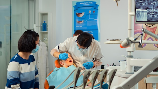 Stomatologist talking with mother of kids, lighting the lamp and examining little girl standing near stomatological chair. Pediatric dentist speaking to woman while nurse preparing sterilized tools.