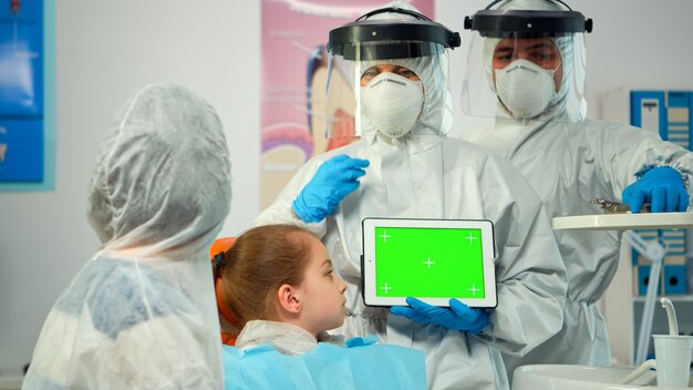 コロナウイルスの流行中にモックアップグリーンスクリーンディスプレイを指しているつなぎ服を着た口腔病学者。グリーンスクリーンモニター付きモニターの使用について説明するクロマキーizolatedクロマPCキーモックアップタッチスクリーン