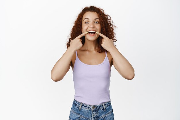 Stomatology, dental clinic. Smiling redhead woman pointing fingers at her white teeth, perfect smile, wearing purple tank top against white background.