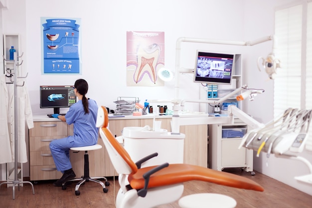 コンピューターで作業している青い制服を着た最新の設備と看護師がいる口腔病学オフィス。