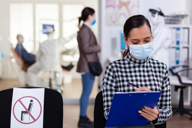 전 세계적으로 발병하는 동안 코로나바이러스 감염에 대한 안전 예방책으로 PPE 옷을 입은 치과 의사와 상담하기 전에 치과 대기 영역을 채우는 구강병 환자.