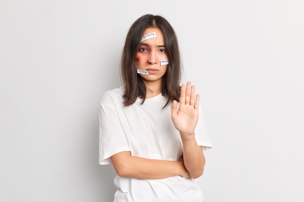 Free photo stop domestic violence. serious unhappy stressed asian woman with traces of beating on face keeps palm forward, has bruise under eye stands indoor. social problems aggression abuse concept