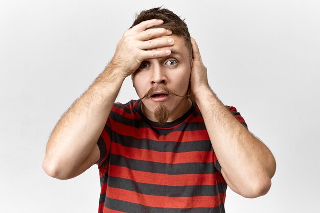 Stop it, this is blowing my mind! Desperate stressed young bearded male being exhausted, having fight with his wife, holding hand on his forehead. Stylish guy looking at camera with panic and anxiety