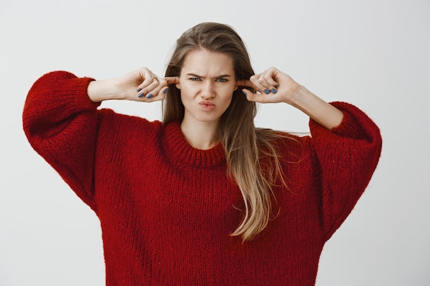 Stop making disturbing noises. Studio shot of displeased annoyed attractive woman in trendy loose sweater, making earplugs with index fingers, covering ears and frowning, hating hear annoying noise