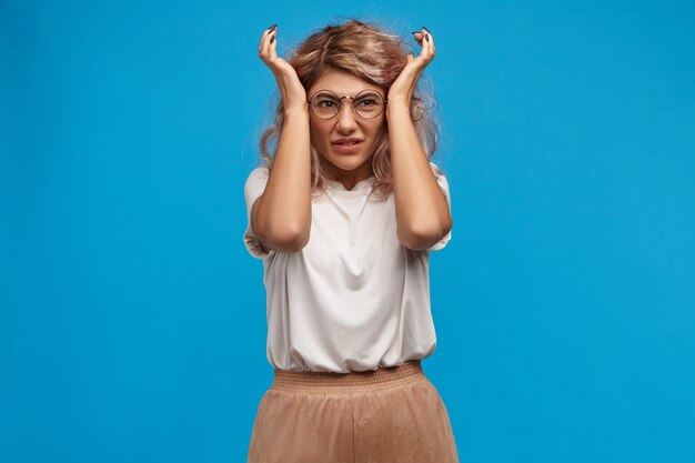 Stop making this irritating noise.  picture of frustrated displeased young female in round eyewear grimacing with pain and squeezing temples or covering ears