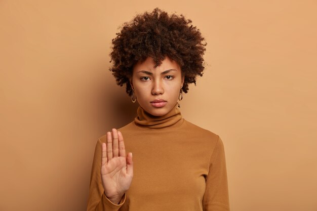 Stop right here. Serious dark skinned woman stands with outstretched hand, makes prohibition gesture, forbids something, smirks face