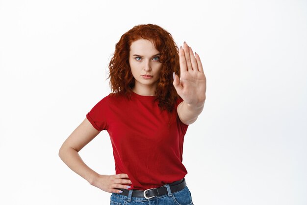 Остановись прямо сейчас. Серьезная и решительная рыжая женщина протягивает руку, чтобы показать жест блокирования, сказать нет, отказаться от чего-то плохого, стоя у белой стены