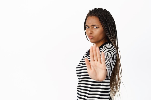 Stop this Serious african woman extending hand at camera showing palm taboo gesture stopping saying no standing over white background