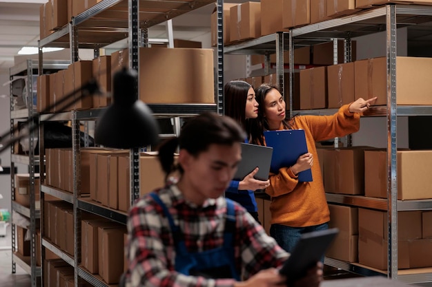 Free photo storehouse asian women employees checking package packing quality in postal storage room. storehouse shipment operator showing goods in stock on shelf to logistics manager