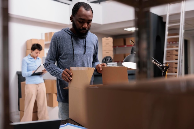 Storehouse supervisor preparing customers orders putting products in cardboard boxes, checking shipping details before delivery packages. Diverse team working in distribution center warehouse