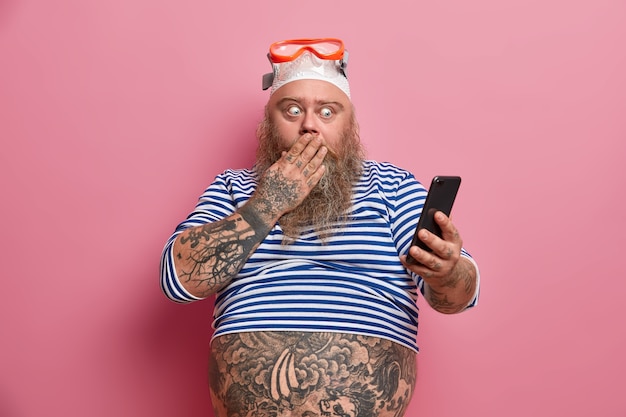 Robusto uomo barbuto scioccato copre la bocca e fissa lo smartphone, legge qualcosa di sorprendente, vestito con camicia da marinaio, occhialini da nuoto, ha la pancia tatuata Foto Gratuite