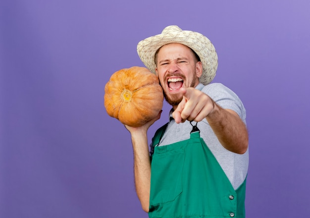 Strained and confident young handsome slavic gardener in uniform and hat holding butternut pumpkin with one hand looking and pointing  isolated on purple wall with copy space