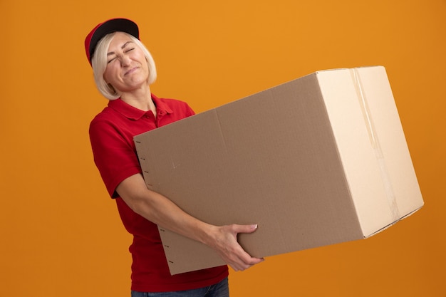 Бесплатное фото Напряженная блондинка средних лет доставщица в красной форме и кепке держит картонную коробку с закрытыми глазами, изолированную на оранжевой стене
