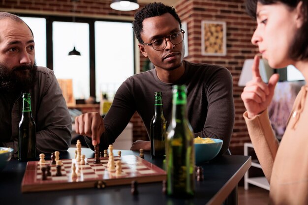 Напряженные многонациональные люди сидят за столом и вместе играют в шахматы. Многорасовые друзья сидят дома в гостиной, наслаждаясь стратегической настольной игрой и закусками.